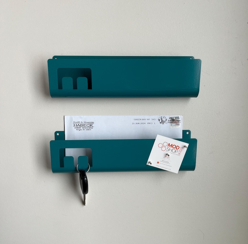 May include: Two teal metal wall-mounted mail organizers with a rectangular cutout on the left side. The bottom organizer holds a white envelope addressed to Scott & Marcia Habeck and a white card with the text Mod Shop Kids and a logo.