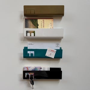 May include: Four rectangular metal wall-mounted organizers in gold, white, teal, and black. Each features a cutout letter E on the left side. The organizers hold various items including a book, envelopes, a business card, and a set of keys.