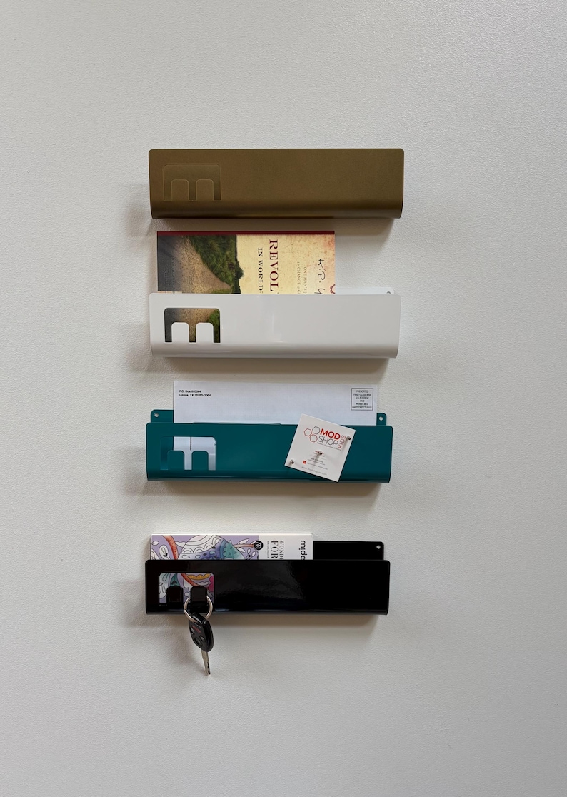 May include: Four rectangular metal wall-mounted organizers in gold, white, teal, and black. Each features a cutout letter E on the left side. The organizers hold various items including a book, envelopes, a business card, and a set of keys.