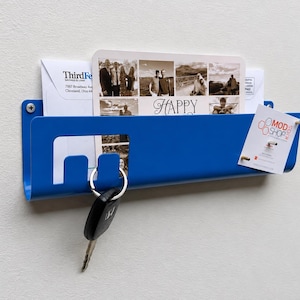 May include: A blue metal wall-mounted mail organizer featuring a rectangular slot for letters and cards, including a photo collage card with the text Happy and a Mod Shop Kids tag. A black car key hangs from a cutout hook on the front edge.