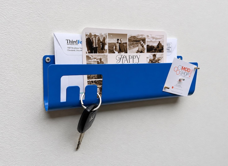 May include: A blue metal wall-mounted mail organizer featuring a rectangular slot for letters and cards, including a photo collage card with the text Happy and a Mod Shop Kids tag. A black car key hangs from a cutout hook on the front edge.