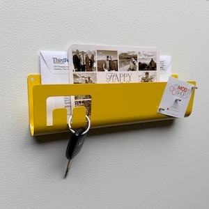 May include: A bright yellow wall-mounted metal mail organizer featuring a rectangular slot for letters and a cutout hook for keys. The organizer holds several envelopes and a small white card with the text MOD SHOP KIDS printed on the front.