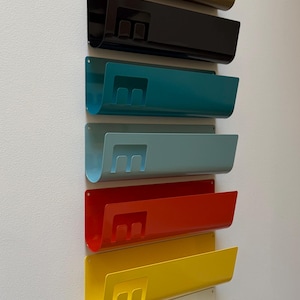 May include: A vertical arrangement of seven wall-mounted metal organizers in olive, black, teal, light blue, red, yellow, and white. Each organizer features a curved U-shaped profile and a cutout letter E on the front face for document storage.