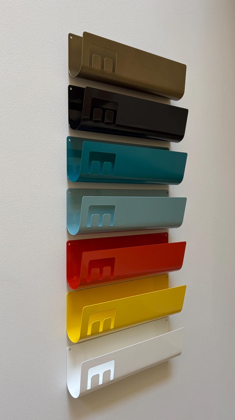 May include: A vertical arrangement of seven wall-mounted metal organizers in olive, black, teal, light blue, red, yellow, and white. Each organizer features a curved U-shaped profile and a cutout letter E on the front face for document storage.