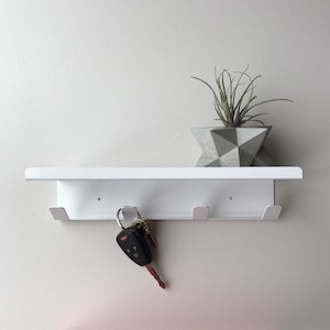 May include: White wall mounted shelf with three hooks and a key ring with keys hanging from one of the hooks. A small potted plant sits on the shelf.