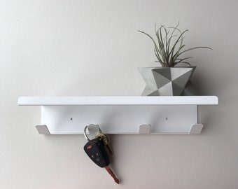 May include: A white metal wall-mounted shelf with three key hooks. A set of keys hangs from one hook. A small succulent plant sits in a geometric gray and white concrete planter on the shelf.