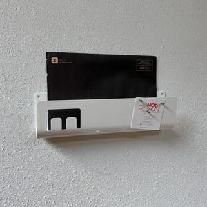 May include: A white wall-mounted metal mail organizer featuring a rectangular cutout design on the left side. A black envelope is tucked inside, and a white business card with the text MOD SHOP KIDS is attached to the right front edge.