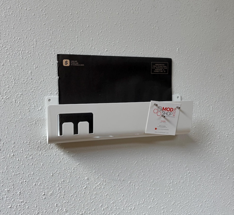 May include: A white wall-mounted metal mail organizer featuring a rectangular cutout design on the left side. A black envelope is tucked inside, and a white business card with the text MOD SHOP KIDS is attached to the right front edge.