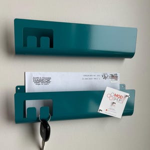 May include: Two teal metal wall-mounted organizers with a rectangular cutout on the left side. The bottom organizer holds a white envelope addressed to Scott & Marcia Habeck and a small white card with the text MOD SHOP KIDS and a logo.