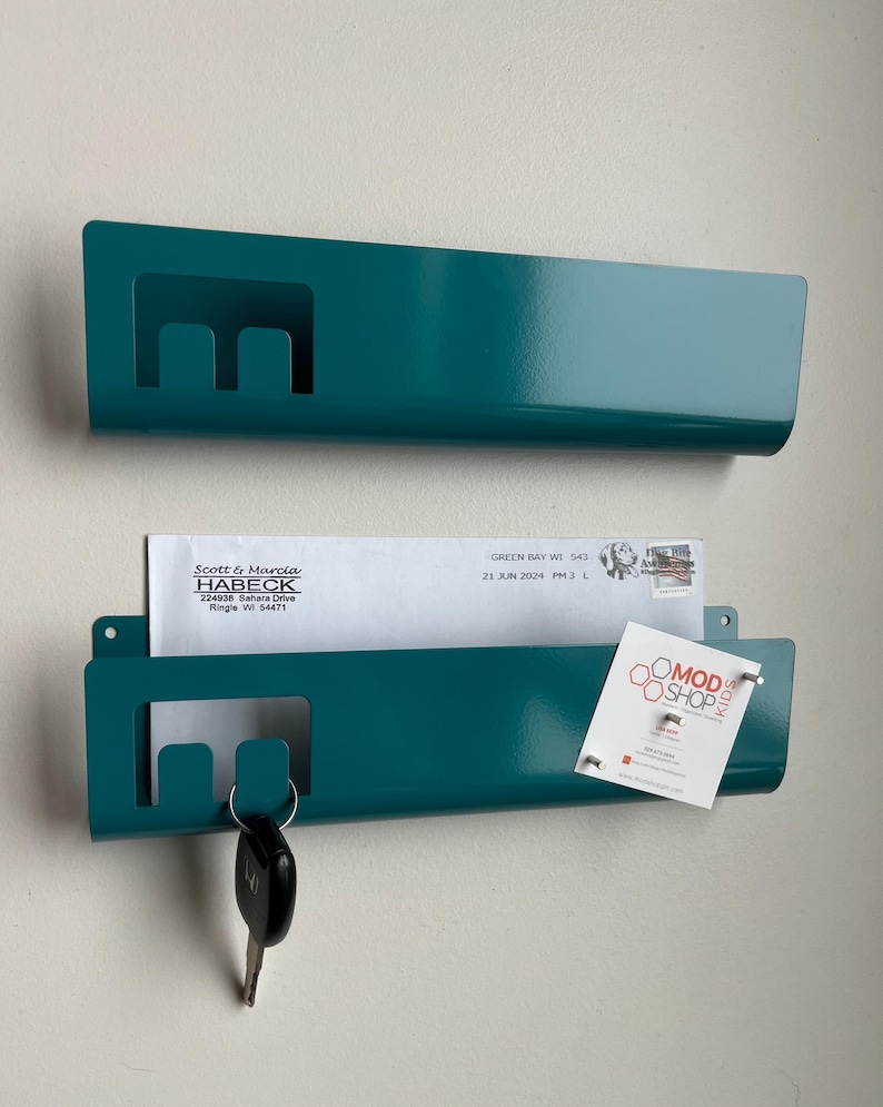 May include: Two teal metal wall-mounted organizers with a rectangular cutout on the left side. The bottom organizer holds a white envelope addressed to Scott & Marcia Habeck and a small white card with the text MOD SHOP KIDS and a logo.