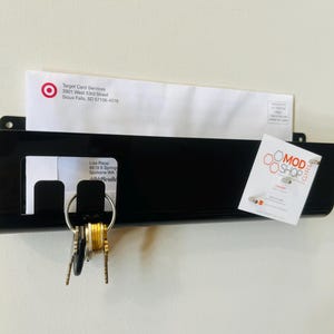 May include: A black wall-mounted mail organizer with a rectangular pocket, featuring a cutout section for hanging keys on a metal ring. The organizer holds a white envelope from Target Card Services and a small white card with the text MOD SHOP GIRL.