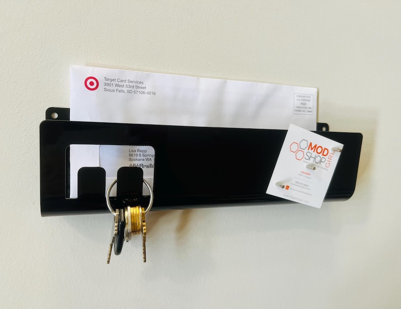 May include: A black wall-mounted mail organizer with a rectangular pocket, featuring a cutout section for hanging keys on a metal ring. The organizer holds a white envelope from Target Card Services and a small white card with the text MOD SHOP GIRL.