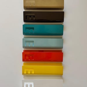 May include: A vertical stack of seven rectangular metal wall-mounted organizers in various colors including gold, black, teal, light blue, red, yellow, and white. Each organizer features a cutout letter E on the left side and a wide open pocket.