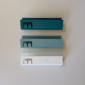 May include: Three rectangular metal wall-mounted organizers arranged vertically. Each unit features a solid back panel with mounting holes, a front shelf, and a cutout section on the left side. The top unit is dark teal, the middle is light blue, and the bottom is white.