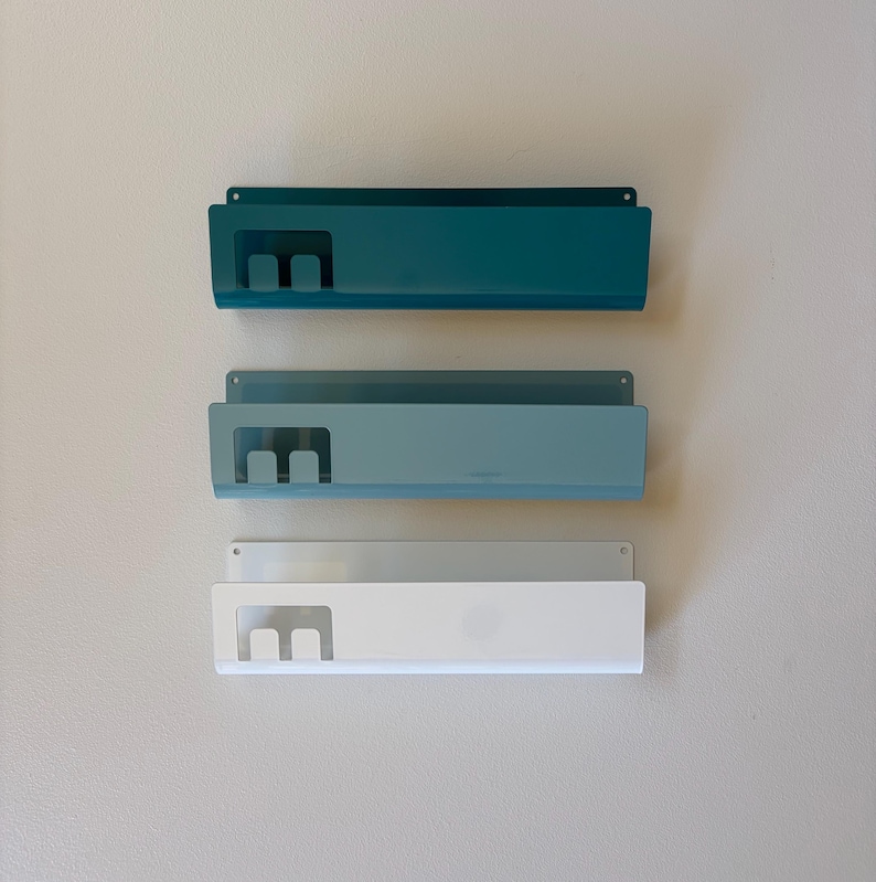 May include: Three rectangular metal wall-mounted organizers arranged vertically. Each unit features a solid back panel with mounting holes, a front shelf, and a cutout section on the left side. The top unit is dark teal, the middle is light blue, and the bottom is white.