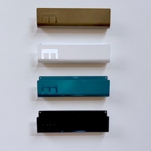 May include: Four rectangular metal wall-mounted storage racks arranged vertically. Each rack features a cutout letter E on the left side. The racks are finished in matte gold, white, teal, and glossy black, each with small mounting holes at the top.
