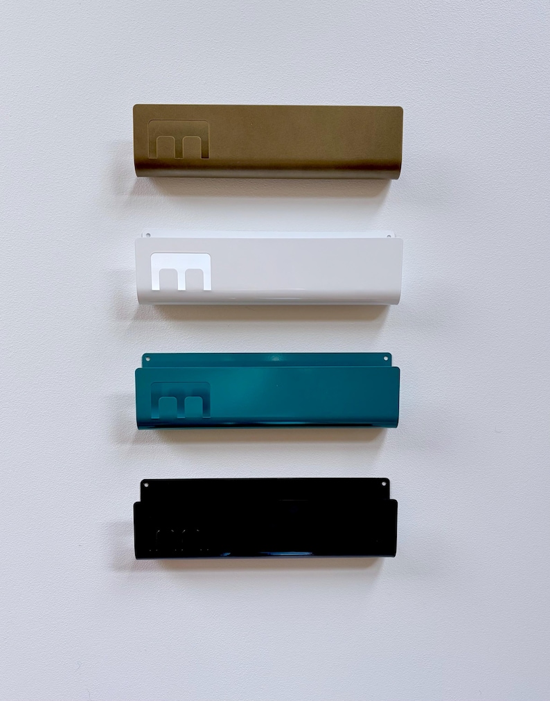 May include: Four rectangular metal wall-mounted storage racks arranged vertically. Each rack features a cutout letter E on the left side. The racks are finished in matte gold, white, teal, and glossy black, each with small mounting holes at the top.