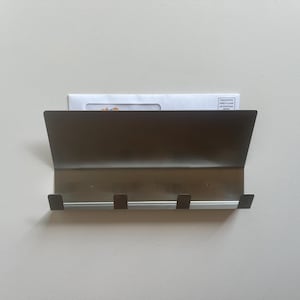 May include: A bronze-colored metal wall-mounted mail organizer with three hooks. The organizer has a sloped top and holds a white envelope with printed text. The hooks are positioned below the mail slot.