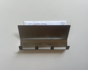May include: A bronze-colored metal wall-mounted mail organizer with three hooks. The organizer has a sloped top and holds a white envelope with printed text. The hooks are positioned below the mail slot.