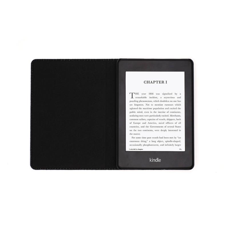 May include: A black Kindle e-reader with a black cover. The screen displays the text "CHAPTER I" and a passage from a book. The Kindle logo is visible at the bottom of the screen.
