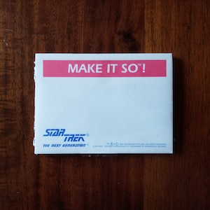 Peut inclure: Un bloc-notes blanc avec une bannière rose en haut qui dit "MAKE IT SO!"  Le bas du bloc-notes porte le texte bleu "STAR TREK" et "THE NEXT GENERATION" en caractères plus petits.