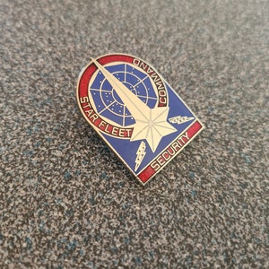 May include: A metal pin with a blue, red, and gold design. The pin features the words "STAR FLEET COMMAND SECURITY" and a starburst design. The pin is in the shape of a shield.