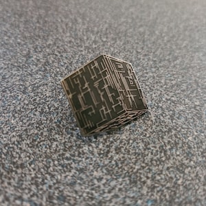 May include: A dark gray, cube-shaped pin with a complex, geometric design etched into its surface. The pin has a metallic appearance and is set against a speckled, gray background. The design features a network of straight lines.
