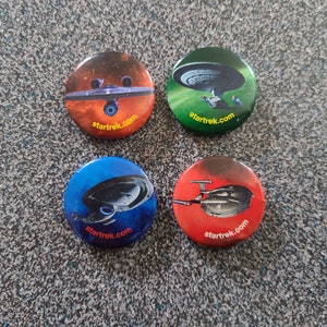 Könnte beinhalten: Vier runde Star Trek-Knöpfe mit verschiedenen Hintergrundfarben und Bildern von Raumschiffen. Die Knöpfe haben alle den Text "startrek.com" aufgedruckt.