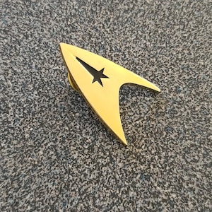 Puede incluir: Un pin dorado con la forma de la insignia de Starfleet de Star Trek. El pin presenta un recorte de estrella negra y un diseño elegante y curvo. El pin está sobre una superficie gris moteada.