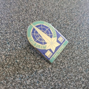 Op de afbeelding: Een gouden Starfleet Command Materiel-speld met een blauwe achtergrond en een groene rand. De speld heeft een gestileerde ster met een radarscherm en een scheeps-pijl.