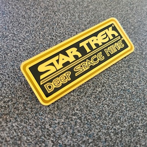 May include: A gold-coloured metal plaque with black lettering that reads "STAR TREK DEEP SPACE NINE".