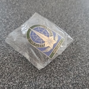 Puede incluir: Un pin de plata con un diseño azul y verde. El pin presenta una estrella con alas y el texto "Star Fleet Command Military".