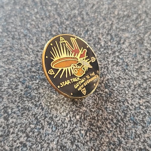 Puede incluir: Un pin de color dorado con fondo negro que presenta una imagen estilizada de la nave espacial Enterprise de Star Trek. El texto "Star Trek Gateway to the New Beginnings" está impreso alrededor de la imagen.