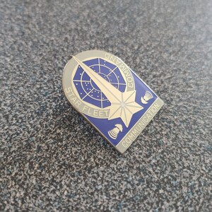 Può includere: Una spilla argentata e blu di Star Fleet Command Communication. La spilla presenta un design a stella con le parole "STAR FLEET COMMAND COMMUNICATION" sui bordi. La spilla è su una superficie strutturata.