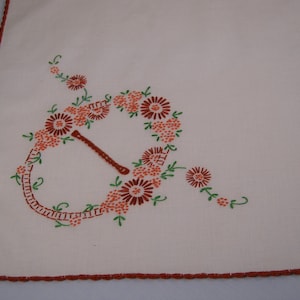 May include: Cream-colored embroidered tablecloth with a floral design. The embroidery features a stylized floral arrangement in shades of orange and brown, with green accents. The edges are finished with a brown, scalloped trim.
