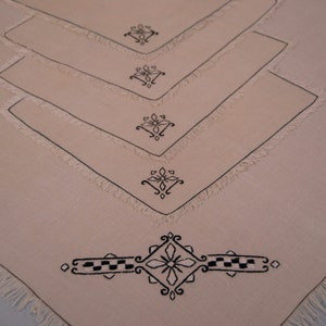 May include: Set of four off-white linen napkins with black embroidered floral designs. The napkins have a fringed edge.