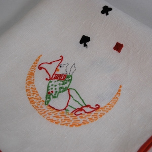Puede incluir: Una toalla de té de lino blanco con ribete rojo y detalles bordados. El bordado presenta un bufón sentado en una luna creciente con acentos naranja y verde. También hay tres símbolos de cartas de juego bordados: un diamante rojo, un trébol negro y un trébol negro.