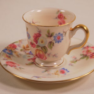 Può includere: Una tazzina da tè e un piattino bianchi con un motivo floreale in tonalità rosa, blu e giallo. La tazza e il piattino hanno un bordo dorato.