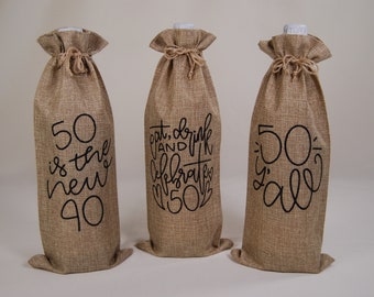 May include: Three brown burlap wine bottle bags with black lettering. The bags have the following text: "50 is the new 40", "Eat, drink and celebrate 50", and "50's y'all".