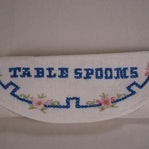 Może przedstawiać: Biały obrus lniany z niebieskim haftem, na którym widnieje napis "TABLE SPOONS" i niebiesko-białą bordurą kwiatową.