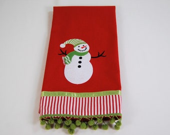 以下が含まれることがあります： 赤いキッチンクロスに白い雪だるまが刺繍されています。雪だるまは緑色の帽子とマフラーをしています。クロスは緑と白のストライプの縁取りと緑のポンポン飾りがあります。