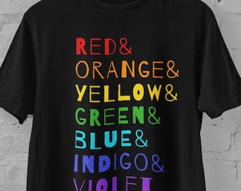 Può includere: Una t-shirt nera a maniche corte con un elenco verticale di testo color arcobaleno che recita RED&, ORANGE&, YELLOW&, GREEN&, BLUE&, INDIGO& e VIOLET. La maglietta è appesa a una gruccia di legno davanti a una parete di mattoni bianchi.