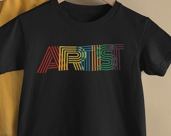 Può includere: Una t-shirt nera a maniche corte con la parola ARTIST stampata in un carattere lineare multicolore di ispirazione retrò. Le lettere sono composte da sottili linee parallele nei toni del rosso, giallo, blu, verde e viola sul petto.