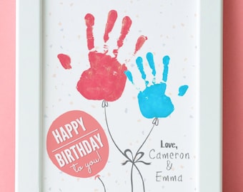 Puede incluir: Una impresión enmarcada en blanco con un diseño de huellas de manos. Dos huellas de manos se representan como globos con cuerdas. El globo rojo con la huella de la mano dice "¡Feliz cumpleaños para ti!" y el globo azul con la huella de la mano tiene un lazo gris. El texto "Love, Cameron & Emma" está escrito debajo de los globos.