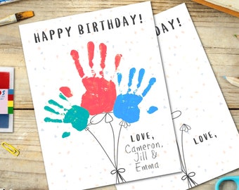 Puede incluir: Una tarjeta de cumpleaños hecha a mano con un mensaje de feliz cumpleaños. La tarjeta presenta tres huellas de manos en rojo, azul y verde, formando una forma de globo. La tarjeta está firmada con los nombres Cameron, Jill y Emma.