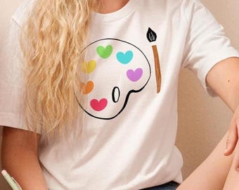 Puede incluir: Camiseta blanca con un diseño de paleta colorida con manchas de pintura en forma de corazón y un pincel. La camiseta se combina con pantalones cortos de mezclilla. Una persona está sentada en una alfombra blanca, sosteniendo un cuaderno y un bolígrafo.