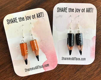 Puede incluir: Dos pares de pendientes de lápiz en tarjetas blancas. Las tarjetas tienen el texto "SHARE the joy of ART!" y "SharemyARTbox.com". Un par de pendientes es negro y el otro es naranja.