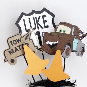 Może przedstawiać: Centralny element dekoracyjny na przyjęcie urodzinowe z brązową lawetą z uśmiechniętą twarzą, tabliczką z napisem "LUKE 1", brązową lawetą z napisem "TOW MATER" oraz dwoma pomarańczowymi stożkami drogowymi. Element dekoracyjny znajduje się w srebrnym wiaderku z czarnym posiekanym papierem.