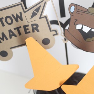 Może przedstawiać: Brązowy kartonowy wycięty obraz ciężar&oacute;wki z napisem "Tow Mater". Na zdjęciu znajdują się dwa pomarańczowe stożki drogowe.