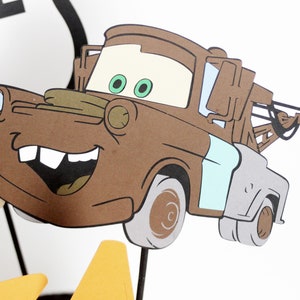 Może przedstawiać: Brązowy kresk&oacute;wkowy samoch&oacute;d holowniczy z niebieskim i szarym panelem bocznym. Samoch&oacute;d ma szeroki uśmiech z zębami i zielone oczy. Samoch&oacute;d holowniczy ma hak na końcu czarnego kabla.